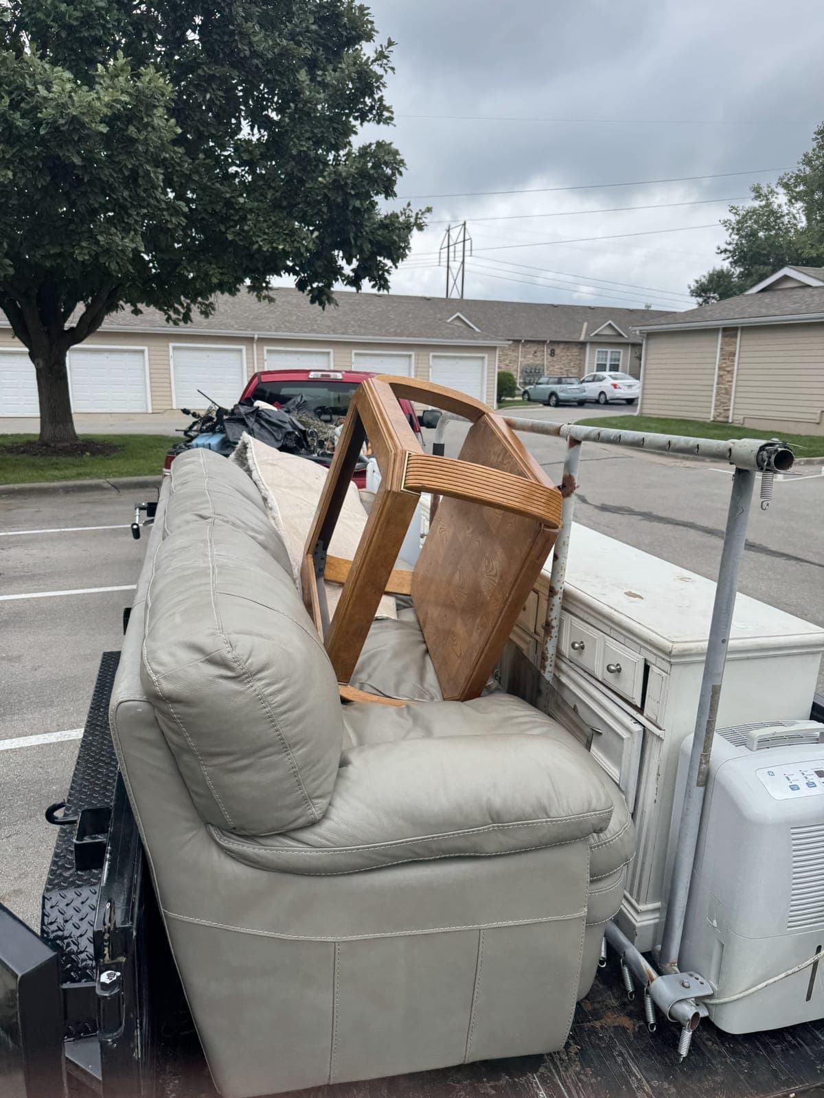 Dump trailer loaded with a light gray sofa, a wooden dining table, a white dresser, and a dehumidifier in an apartment complex parking lot on a furniture-removal job in the Kansas City metro