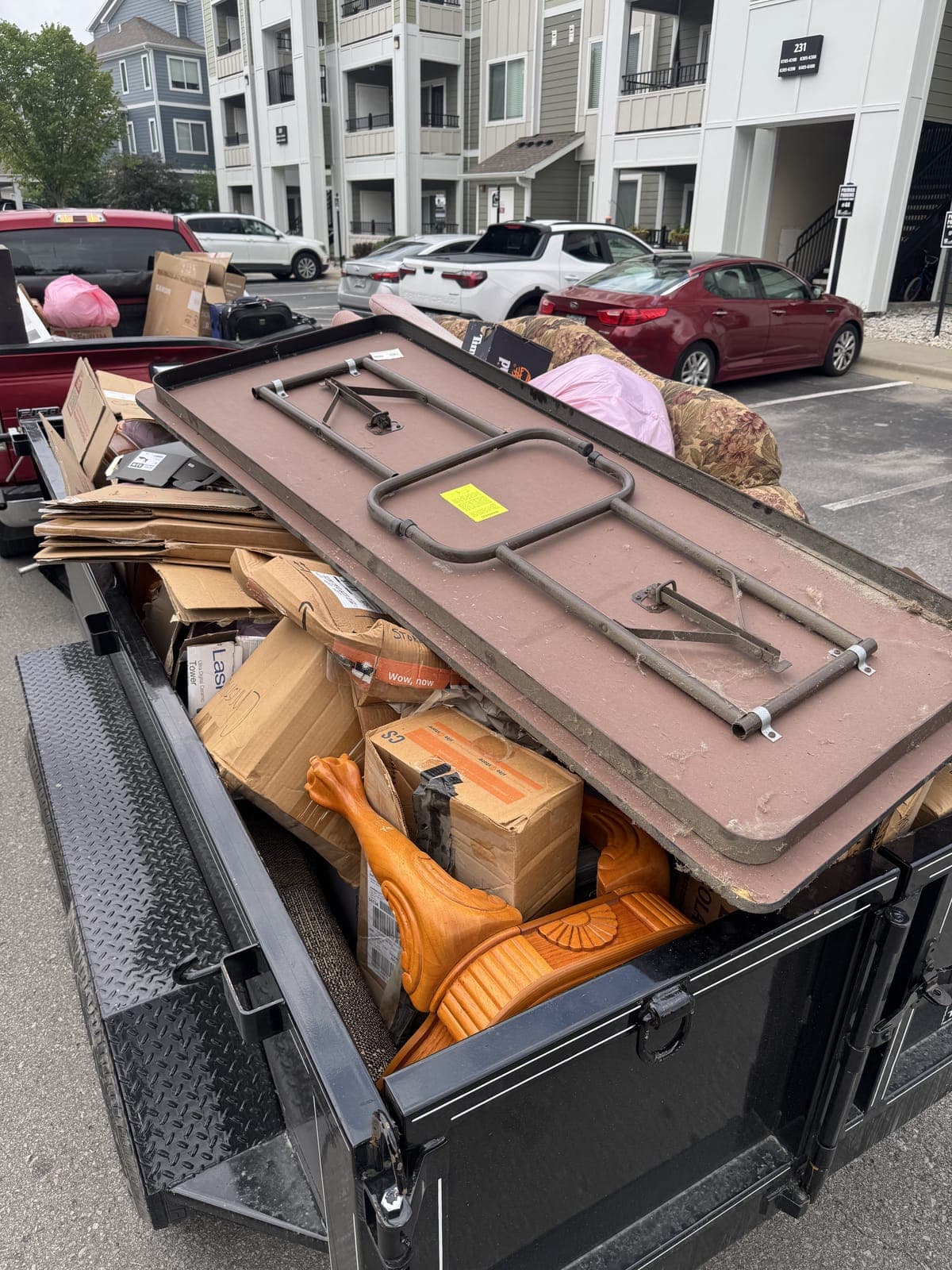 Dump trailer loaded in an apartment complex parking lot with a folding table, moving boxes, and leftover furniture from a tenant move-out cleanout in the Kansas City metro