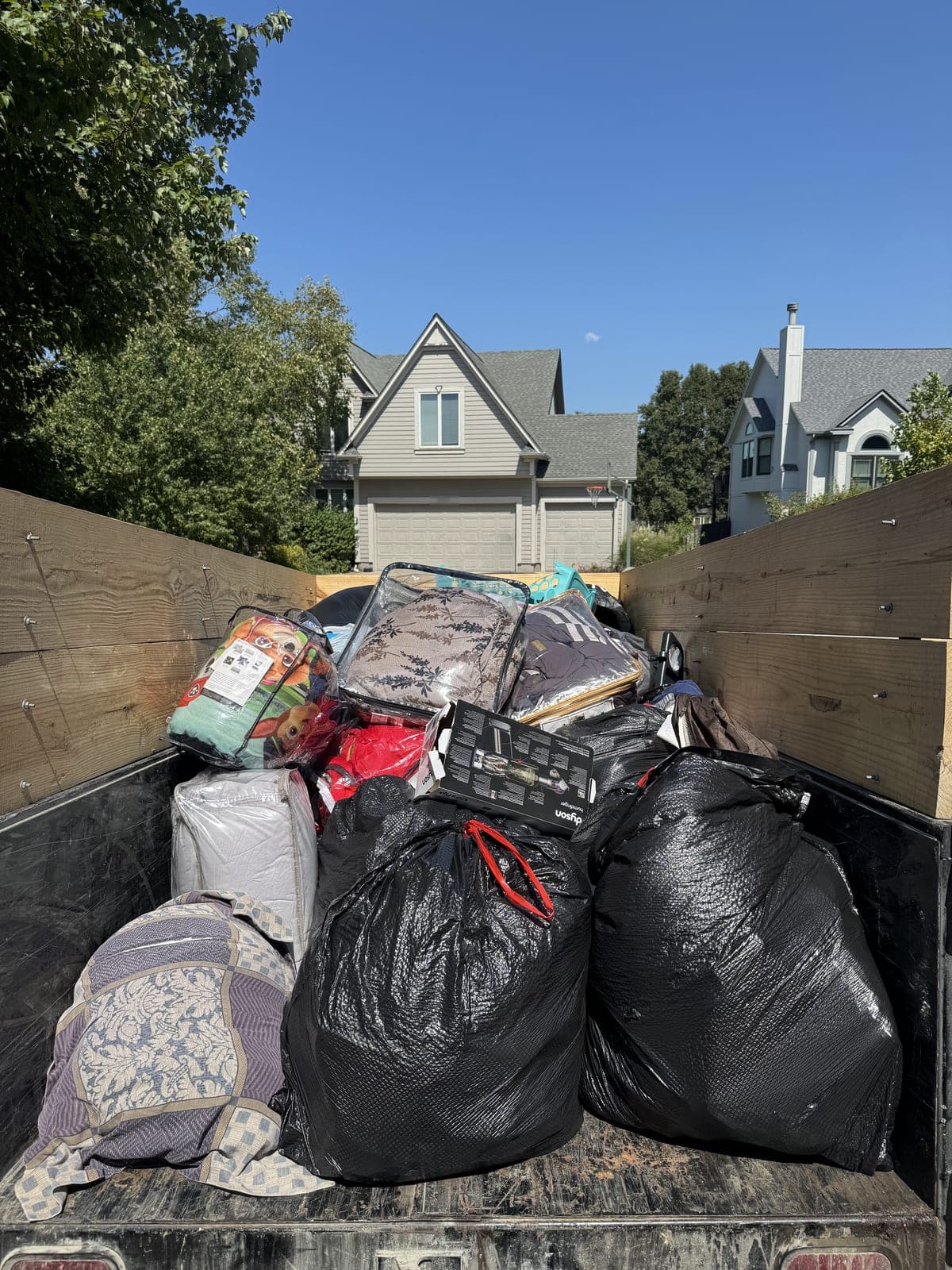Dump trailer with raised wood side extensions loaded curbside in a residential neighborhood with large tied trash bags, packaged comforters and bedding, and a folded quilt on a household cleanout in the Kansas City metro