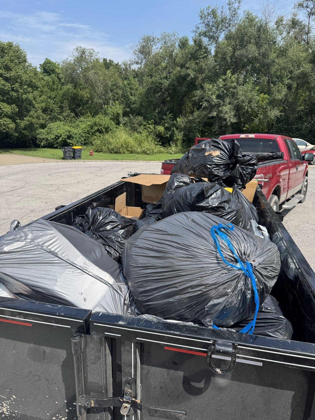 Dump trailer mounded above the rails with tied black bags and flattened cardboard, with a loaded pickup truck parked behind it on a Kansas City metro cleanout job
