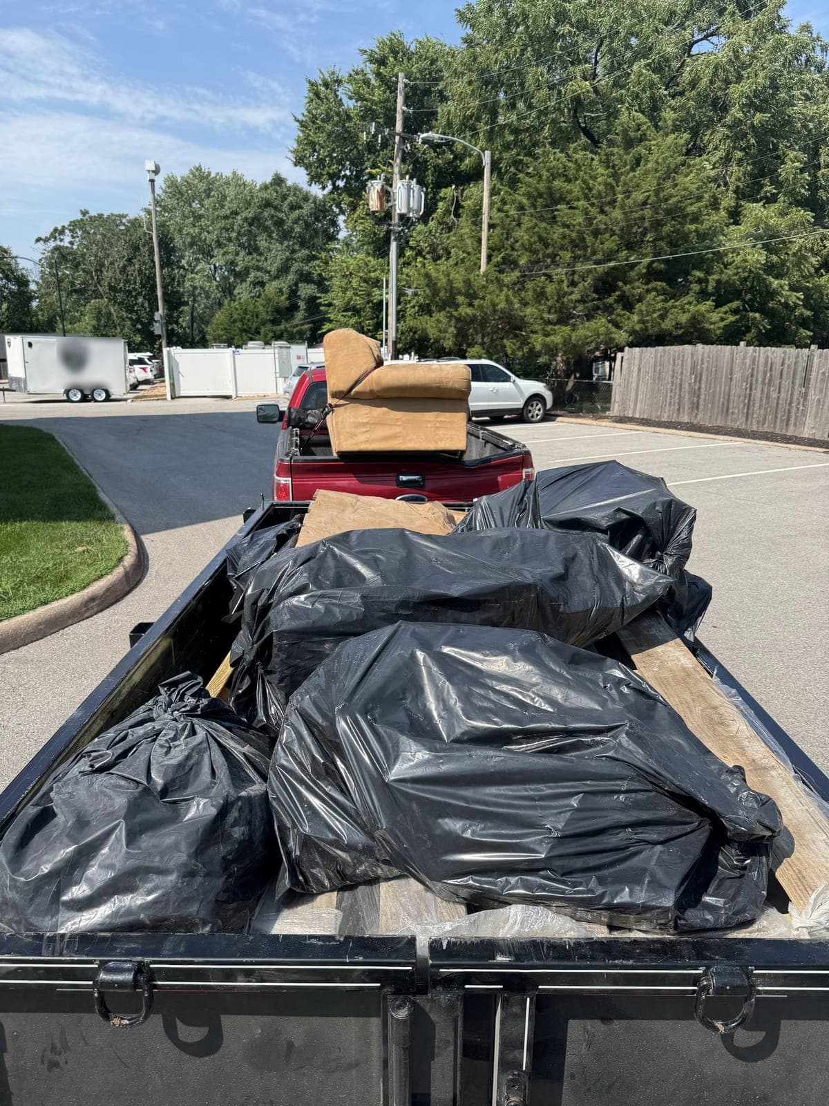 Dump trailer filled with bagged debris and lumber, with an upholstered couch loaded in the pickup truck ahead of it, on a junk-removal job in a Kansas City metro parking lot