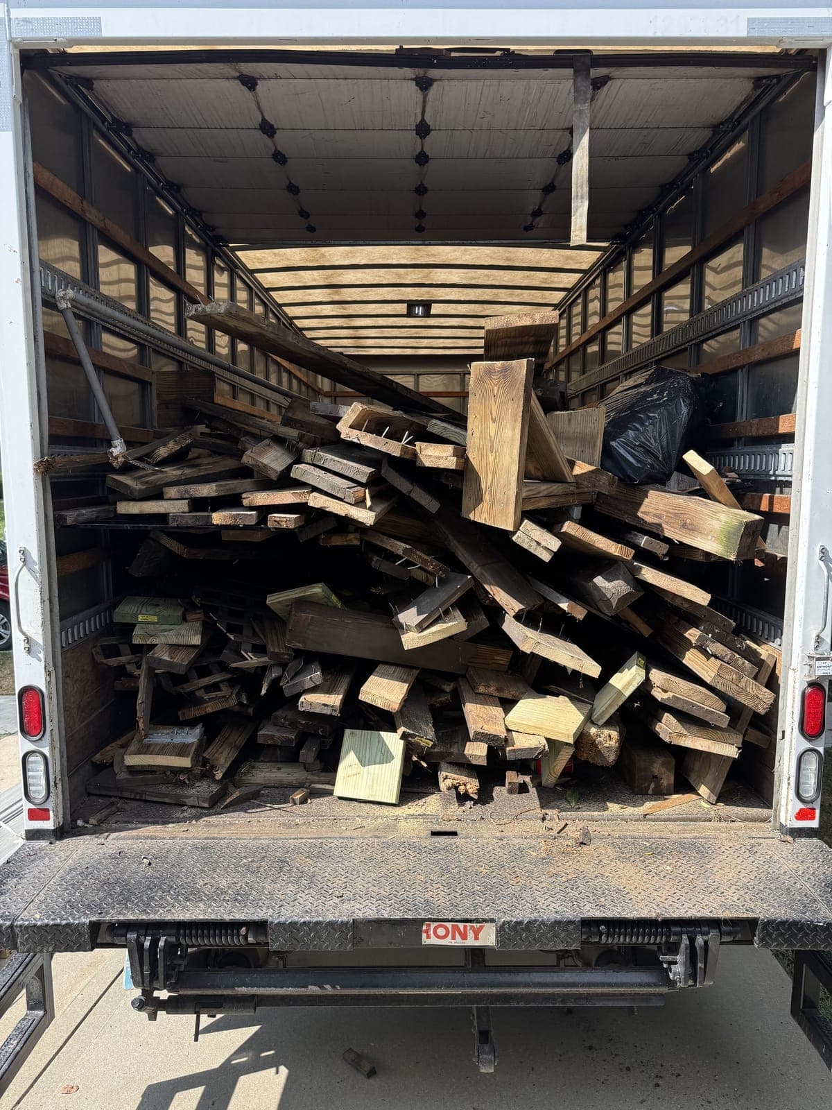 Rear view of a 26-foot box truck with the liftgate down, filled shoulder-high with torn-out deck boards and framing lumber on a construction debris haul in the Kansas City metro
