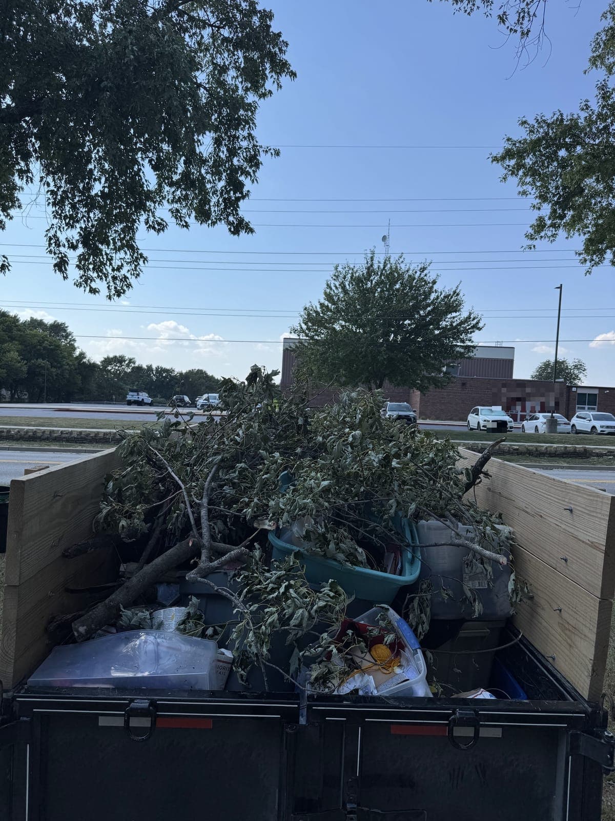 Dump trailer with raised wood side extensions loaded with cut tree branches and brush over plastic storage tubs and bagged household junk, parked on a residential street in the Kansas City metro