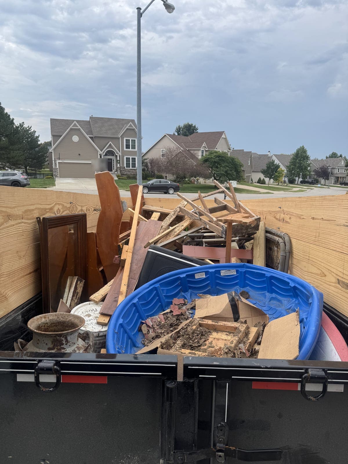 Dump trailer with raised wood side extensions loaded curbside in a residential neighborhood with broken lumber, a cabinet door, a wooden chair frame, a kiddie pool of debris, and a rusted planter on a cleanout job in the Kansas City metro