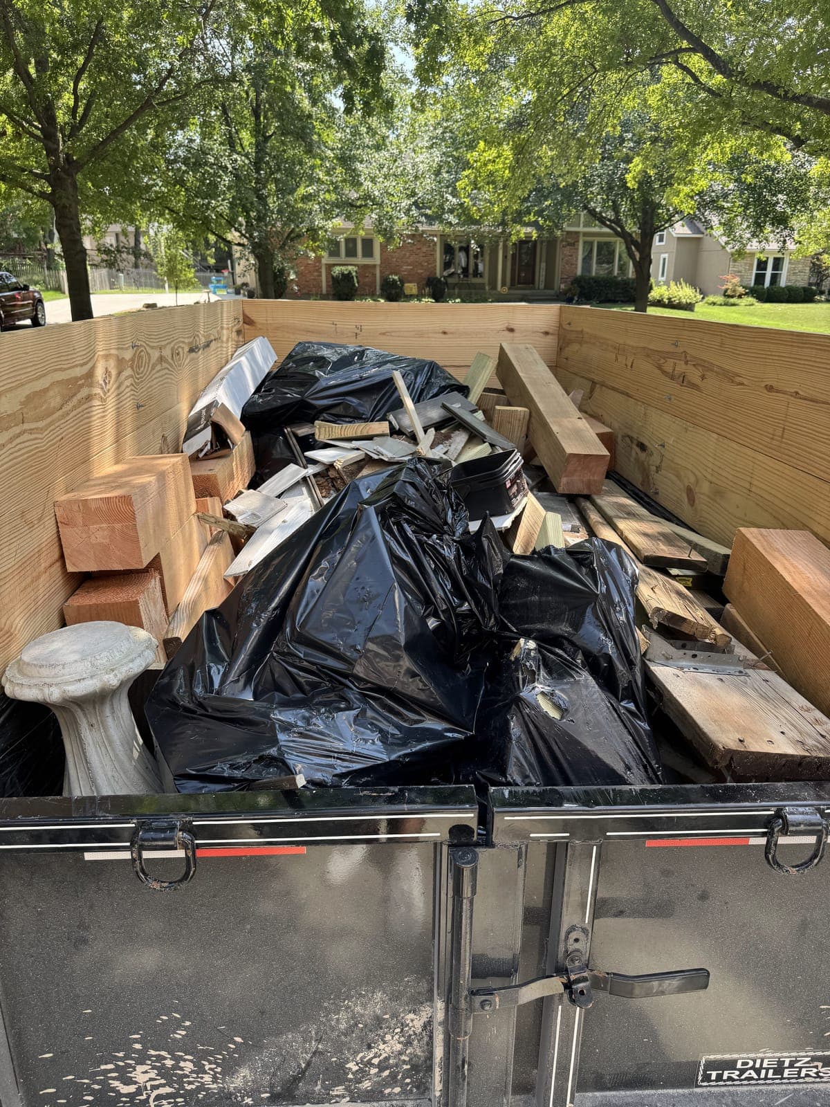 Dump trailer with raised wood side extensions loaded curbside with dimensional lumber, tied black debris bags, and a concrete pedestal on a construction debris haul in the Kansas City metro