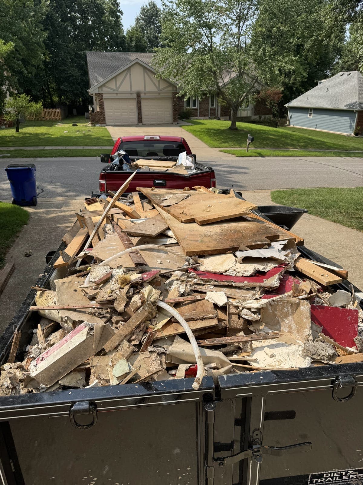 Dump trailer and pickup truck loaded curbside with broken drywall, dimensional lumber, plywood sheets, and trim board on a construction debris haul in the Kansas City metro