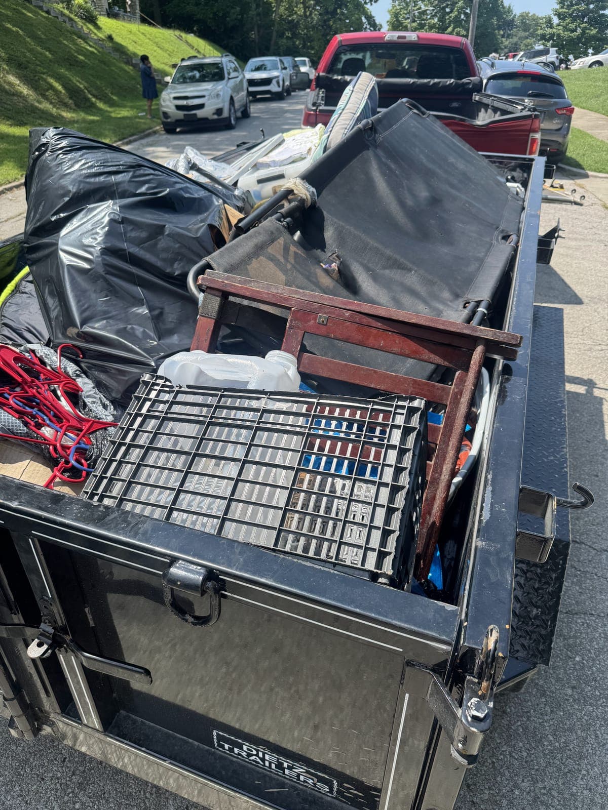 Dump trailer and pickup truck loaded on a residential street with a folding cot, a wooden chair, storage crates, and bagged household junk on a junk-removal job in the Kansas City metro