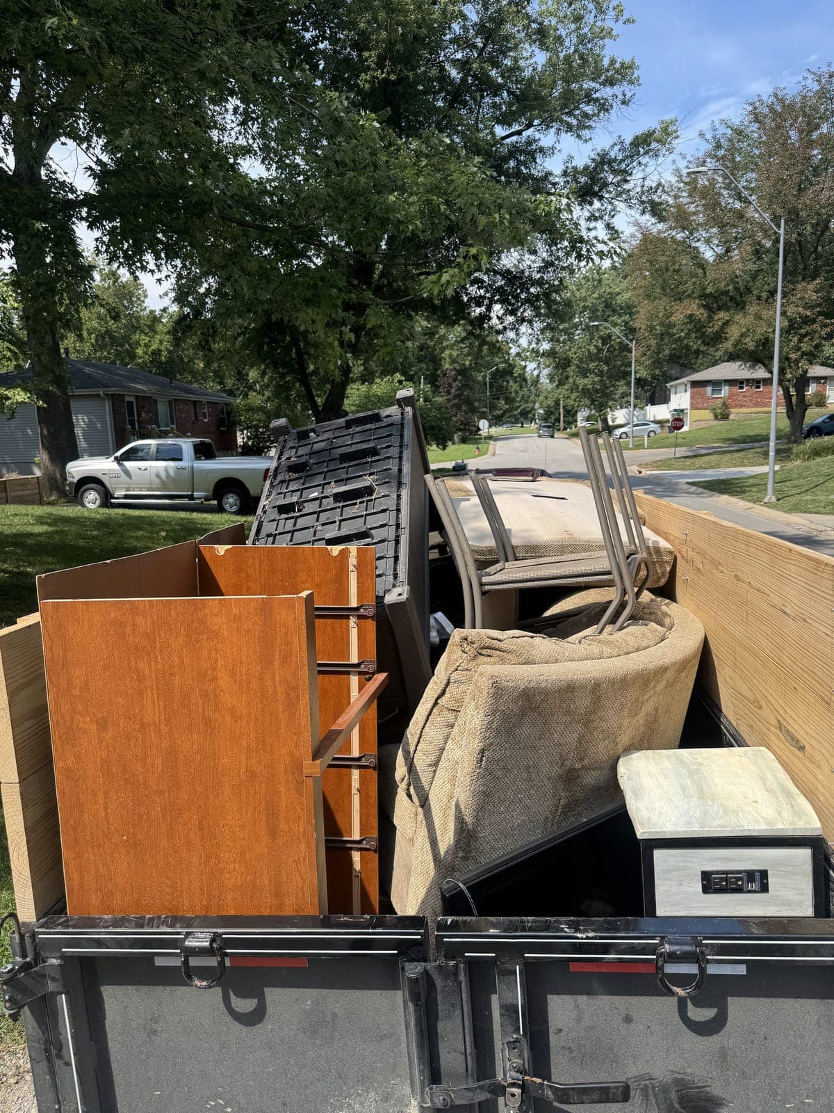 Dump trailer with raised wood side extensions loaded curbside with dresser panels, a mattress, stacked patio chairs, and a black storage crate on a furniture removal job in the Kansas City metro
