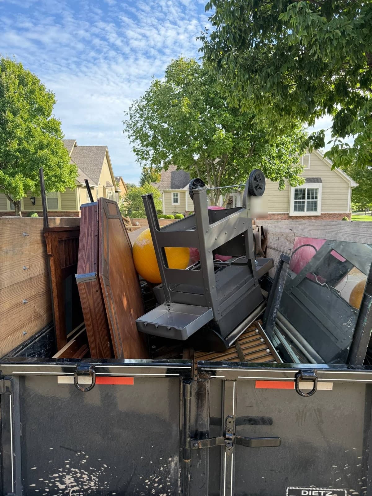 Dump trailer with raised wood side extensions loaded on a residential driveway with an upturned grey steel utility wagon, a large yellow exercise ball, wooden headboard and table panels, bed slats and a dark glass panel on a garage cleanout in the Kansas City metro