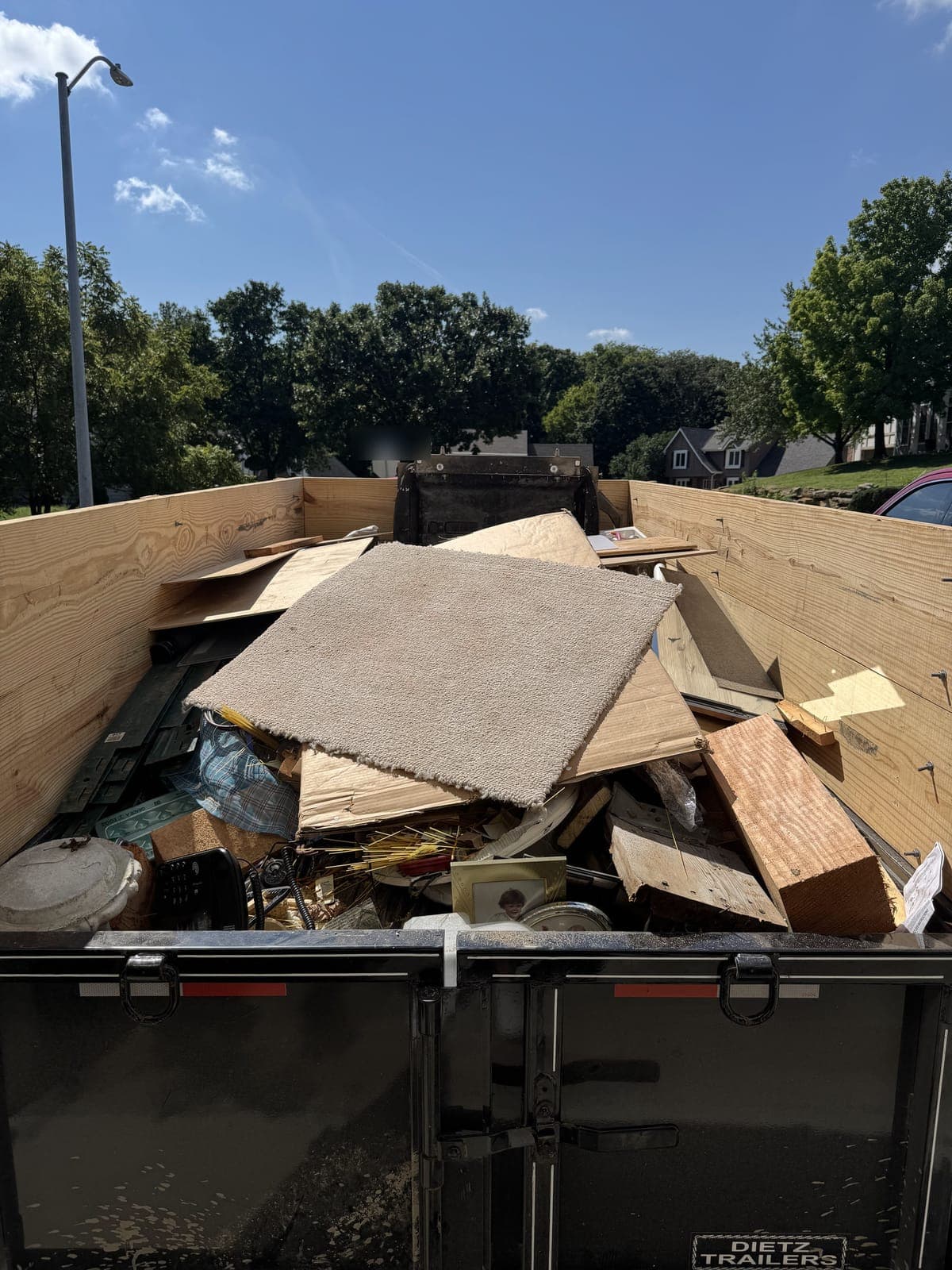 Dump trailer with raised wood side extensions loaded with carpet remnants, plywood, dimensional lumber, and mixed household items on a cleanout job in the Kansas City metro