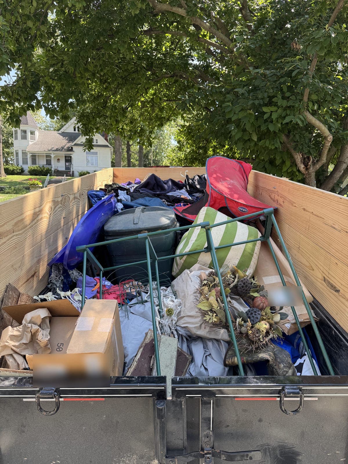 Dump trailer with raised wood side extensions loaded with storage bins, folded clothing, a pet carrier, cardboard boxes, and a dried floral arrangement on a household cleanout in the Kansas City metro