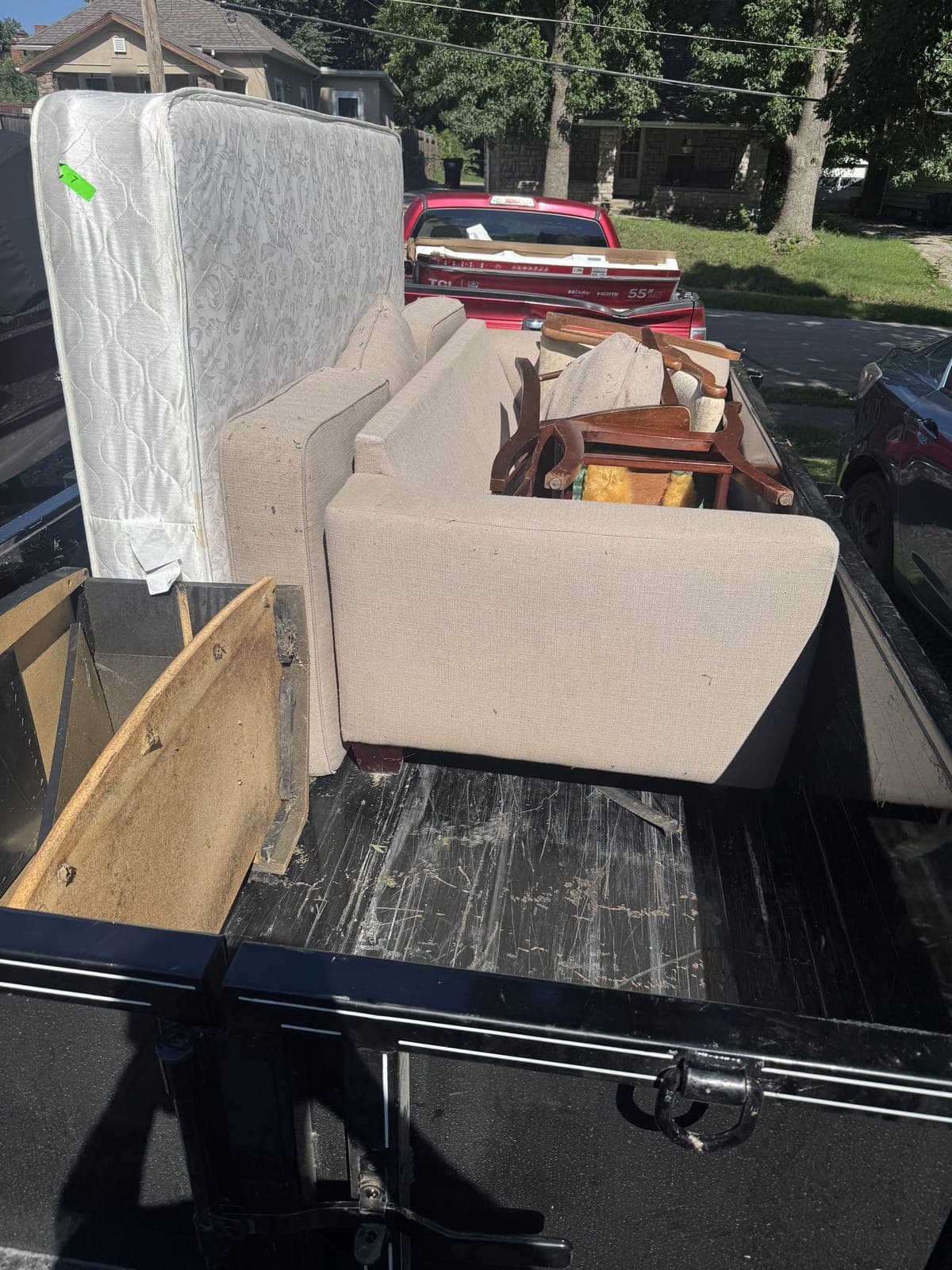 Dump trailer loaded curbside with a mattress, an upholstered sofa, wooden dining chairs, and a headboard on a residential mattress and furniture haul in the Kansas City metro