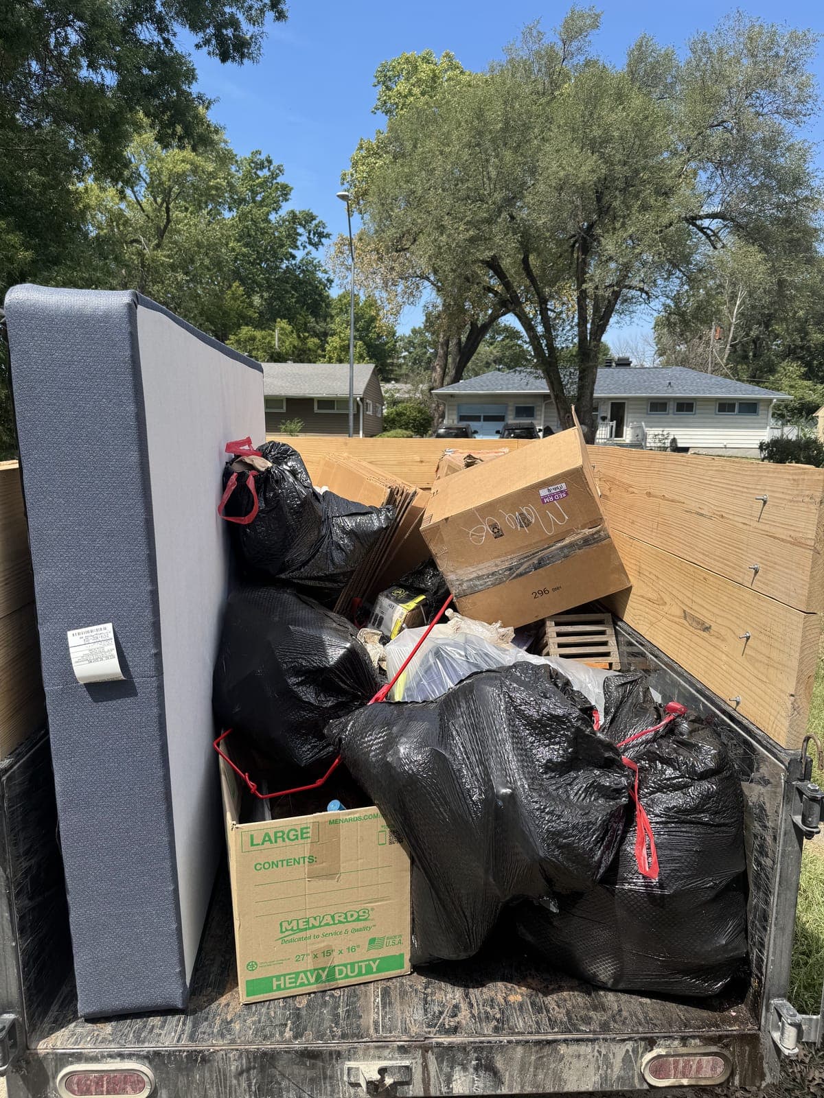 Dump trailer with raised wood side extensions loaded curbside in a residential neighborhood with a mattress standing on edge, tied black trash bags, flattened cardboard, and labeled moving boxes on a household cleanout in the Kansas City metro