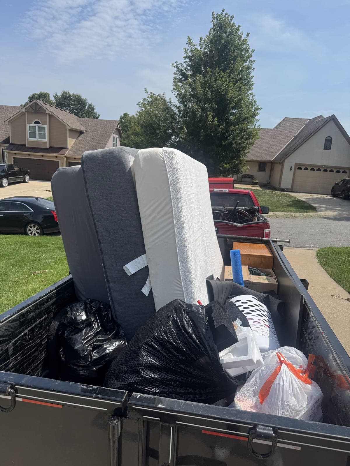 Dump trailer loaded curbside in a residential neighborhood with a mattress, a box spring, sofa cushions, a drawer unit, and bagged household items on a cleanout job in the Kansas City metro