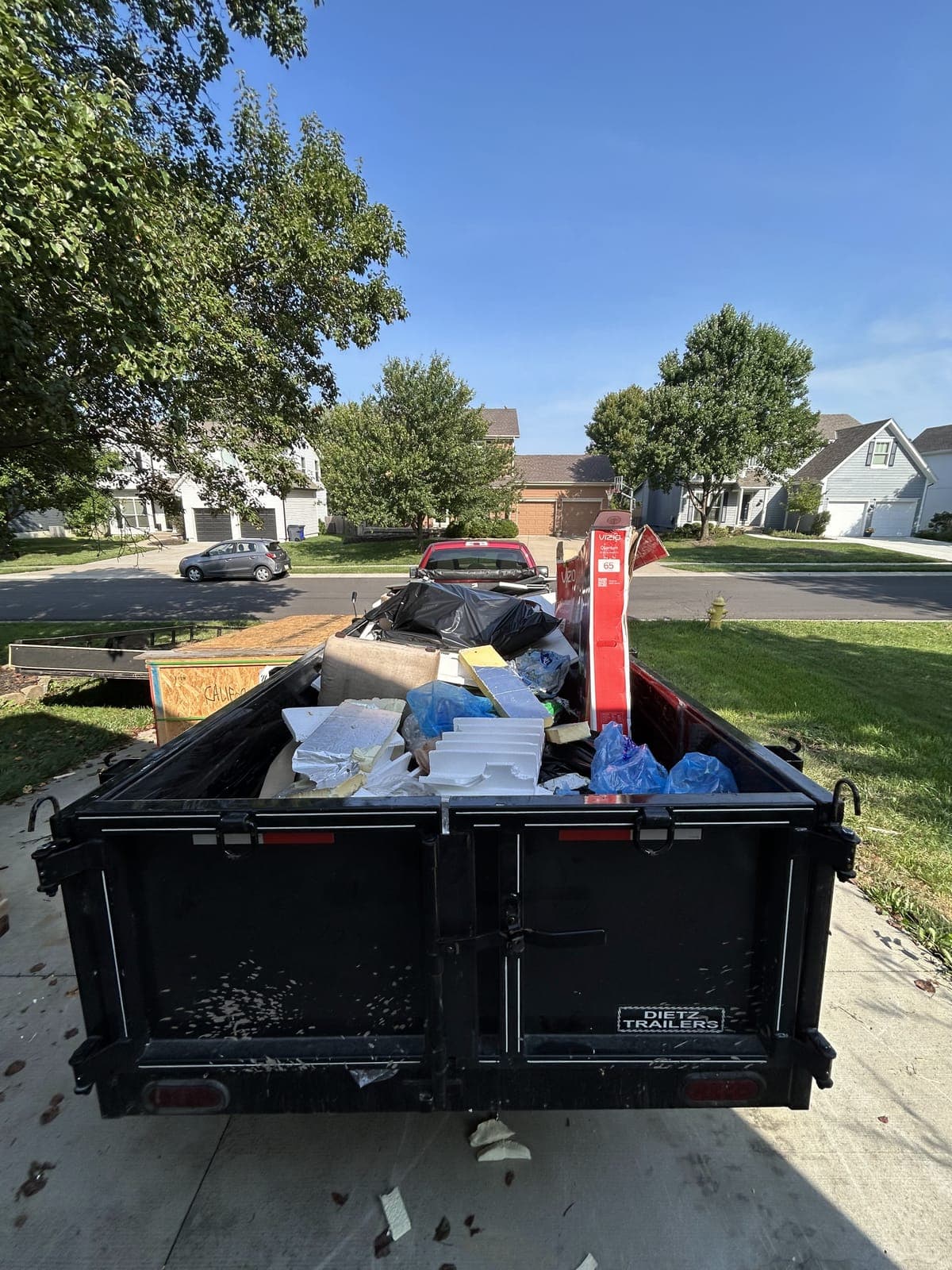 Dump trailer parked on a driveway loaded with bagged household items, foam packaging, cardboard, and a step ladder on a cleanout job in the Kansas City metro