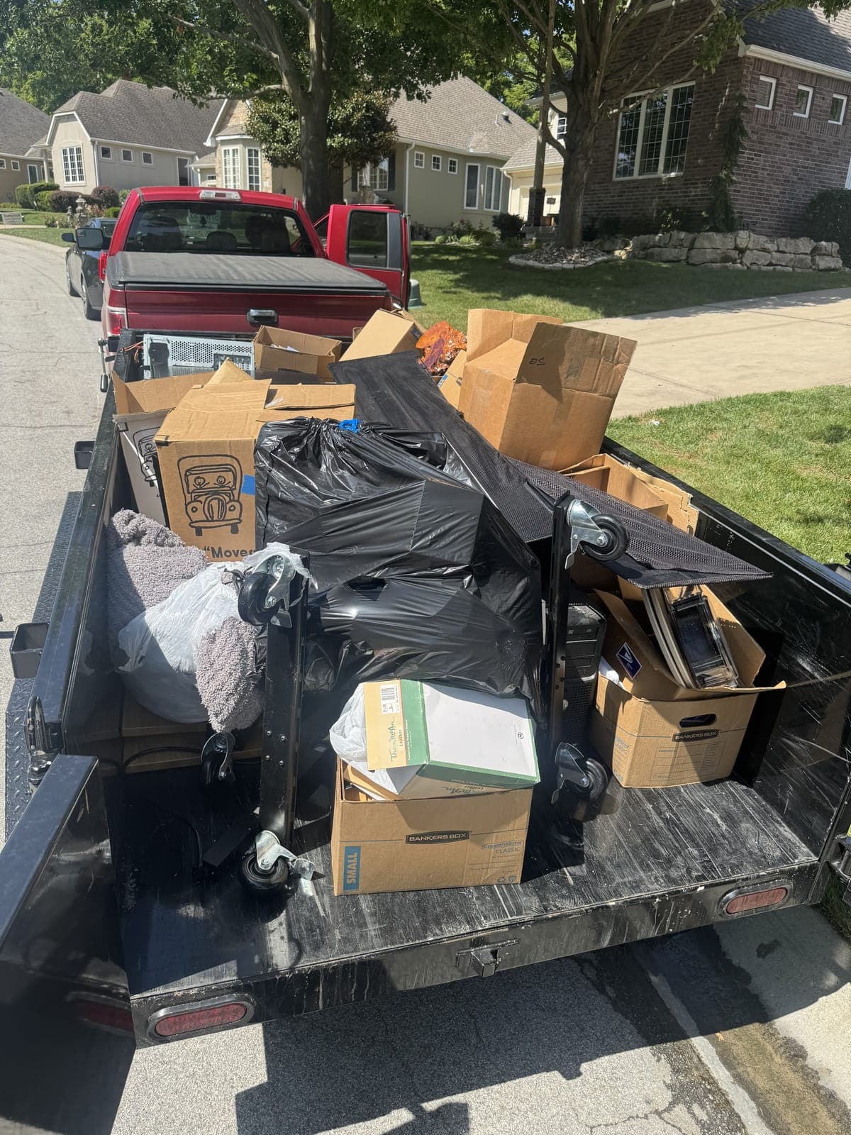 Dump trailer loaded curbside with moving boxes, bagged household items, rolled carpet, and an office chair on a residential move-out cleanout in the Kansas City metro