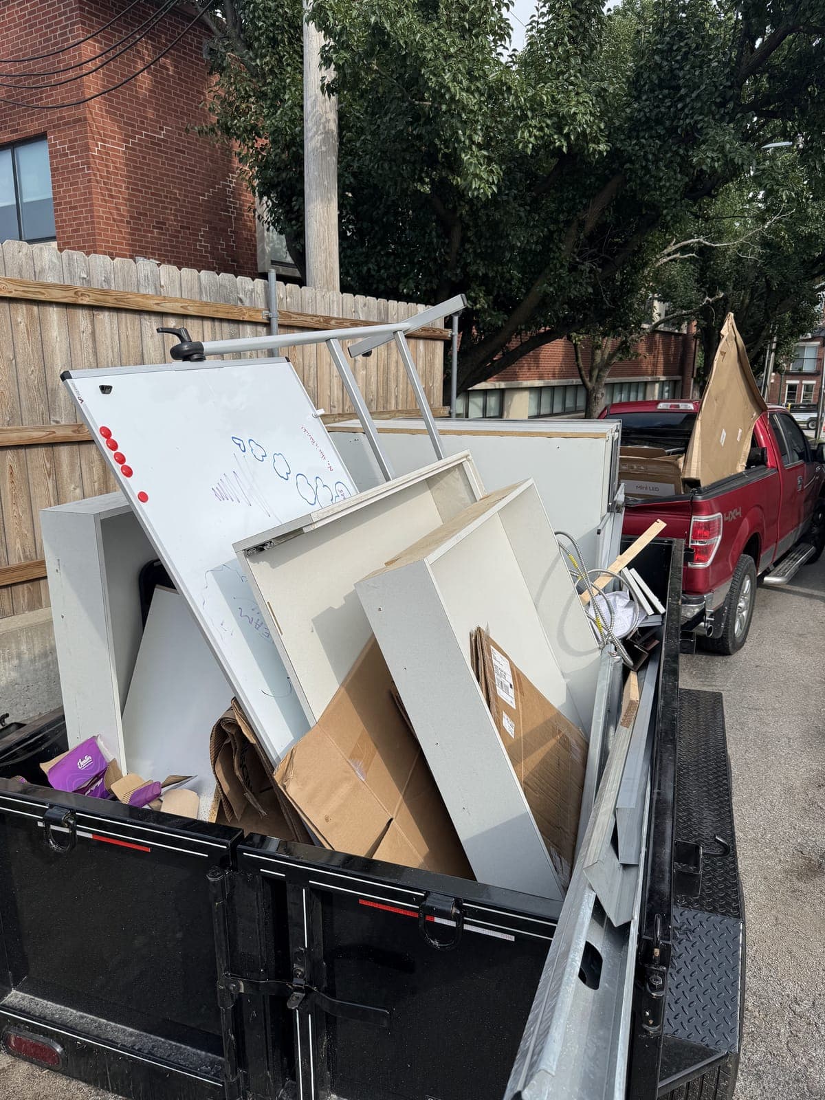 Dump trailer loaded curbside with a whiteboard, office cabinets, shelving, and flattened packing boxes on a commercial office cleanout in the Kansas City metro