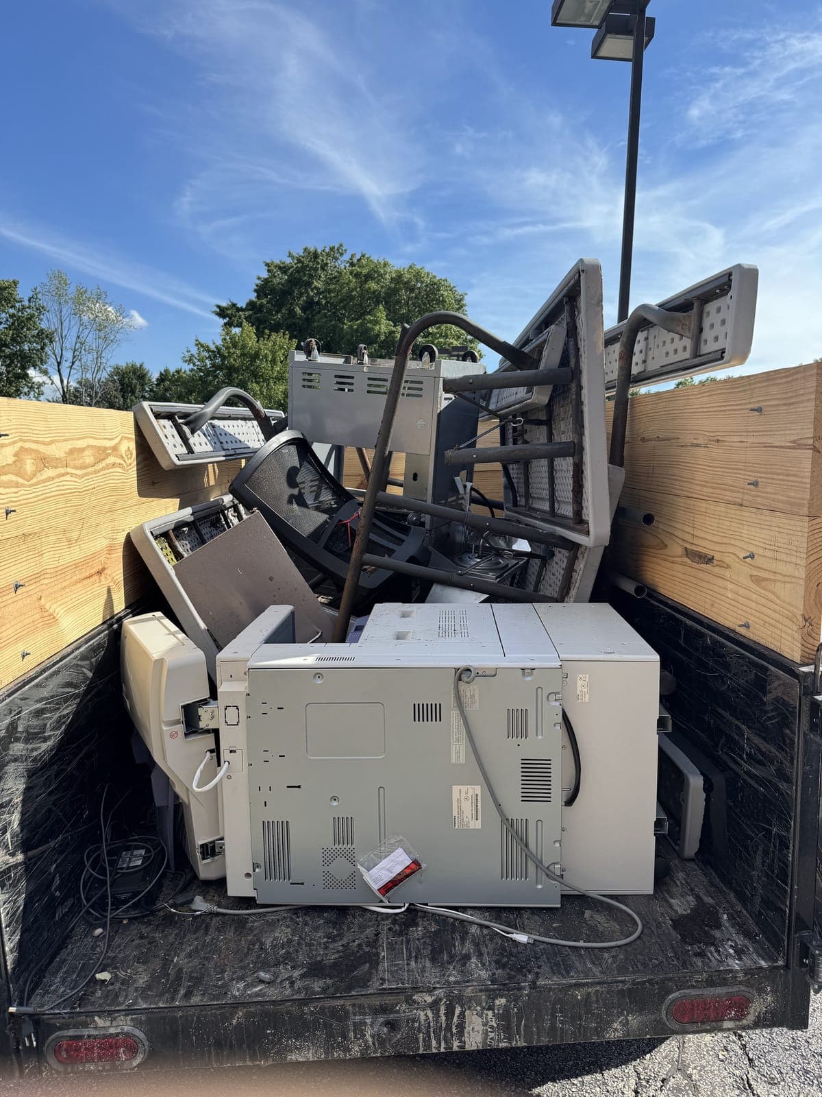 Dump trailer with raised wood side extensions loaded with office copiers, printers, stacked folding tables, a mesh office chair, and a treadmill on a commercial equipment haul in the Kansas City metro