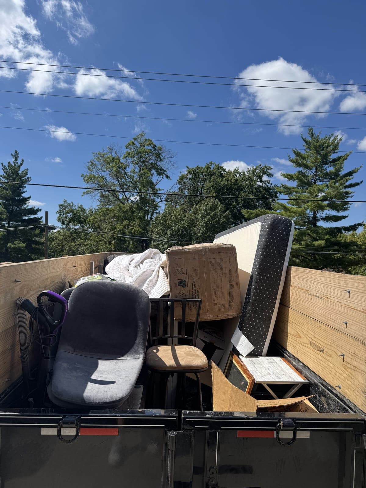A dump trailer with wooden side boards loaded with an office chair, a mattress, a bar stool, a vacuum, a small end table and cardboard boxes on a household junk haul in the Kansas City metro