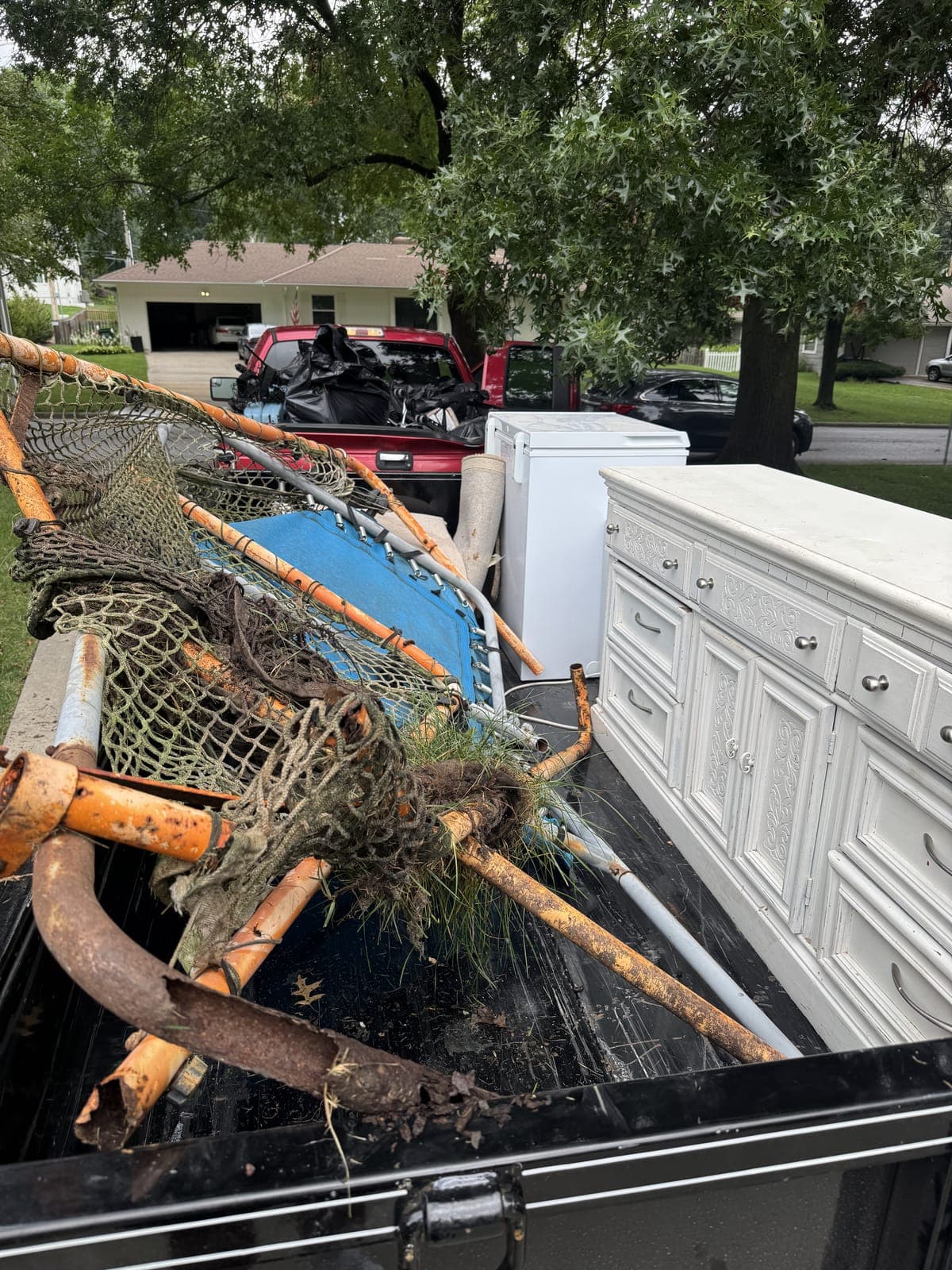 Dump trailer loaded with a dismantled trampoline frame and safety netting, a chest freezer, and a white dresser, with a loaded pickup truck behind it on a residential junk-removal job in the Kansas City metro