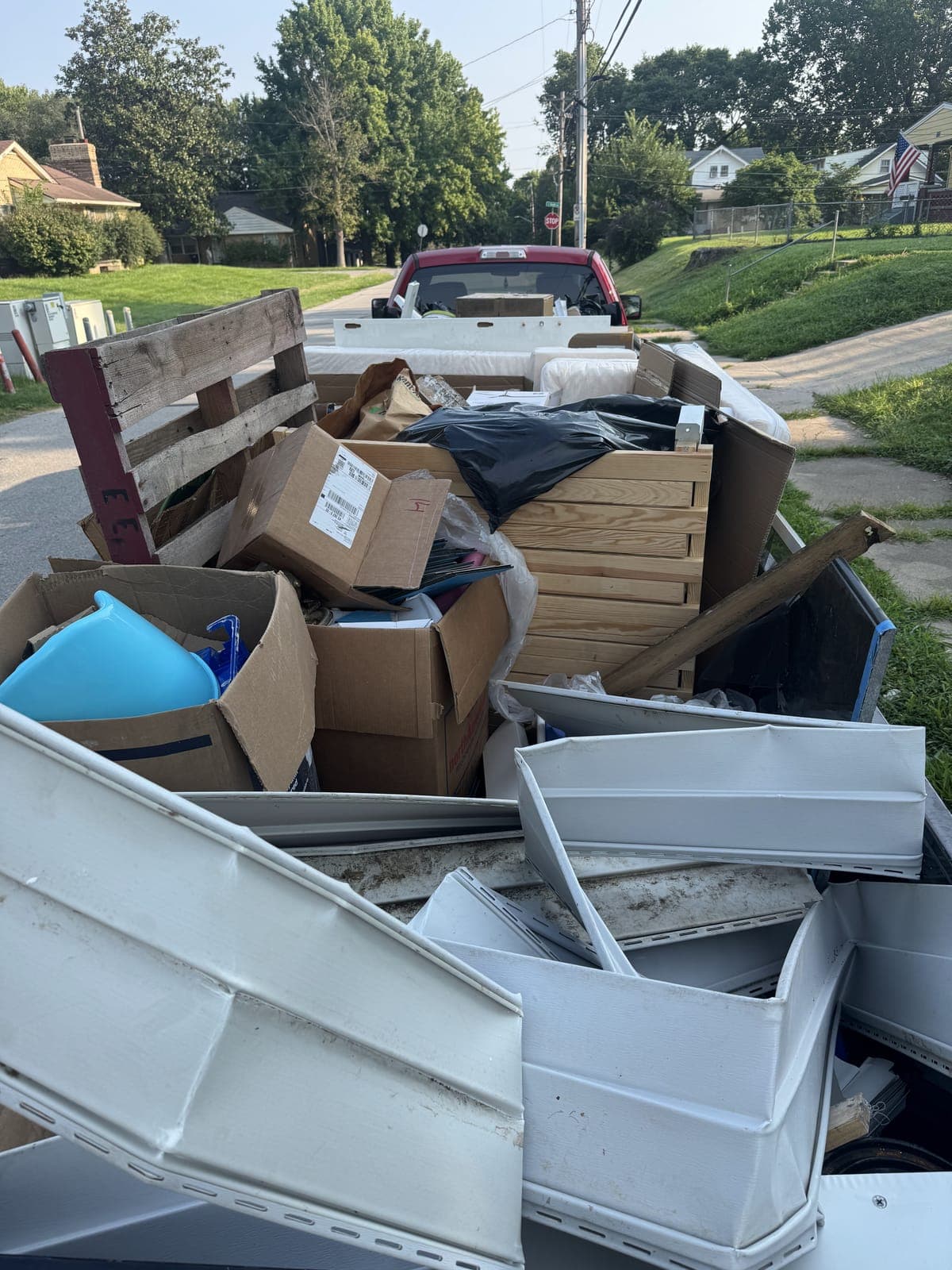 Dump trailer and pickup truck loaded on a residential street with cardboard boxes, bagged junk, a wooden crate, a blue plastic tub, and stacked white panels on a junk-removal job in the Kansas City metro