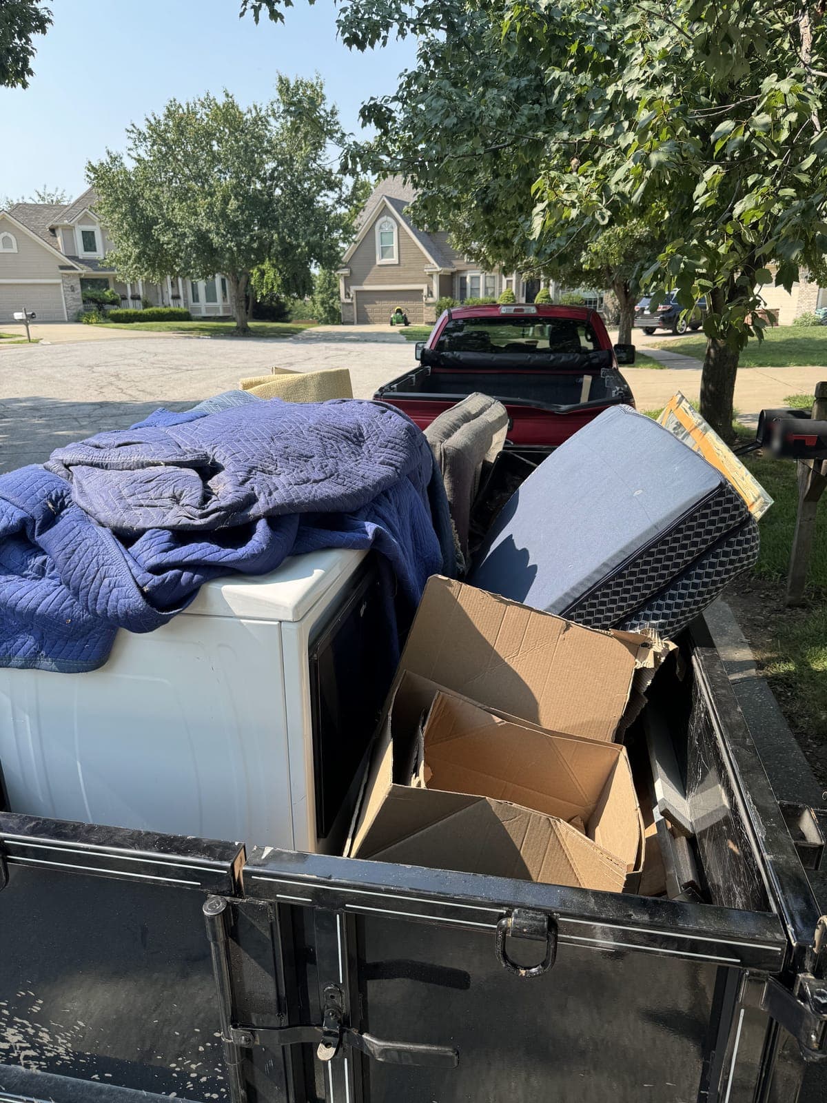 Dump trailer loaded on a residential street with a mattress, a white appliance, flattened cardboard boxes, moving blankets, and a framed picture on a junk-removal job in the Kansas City metro