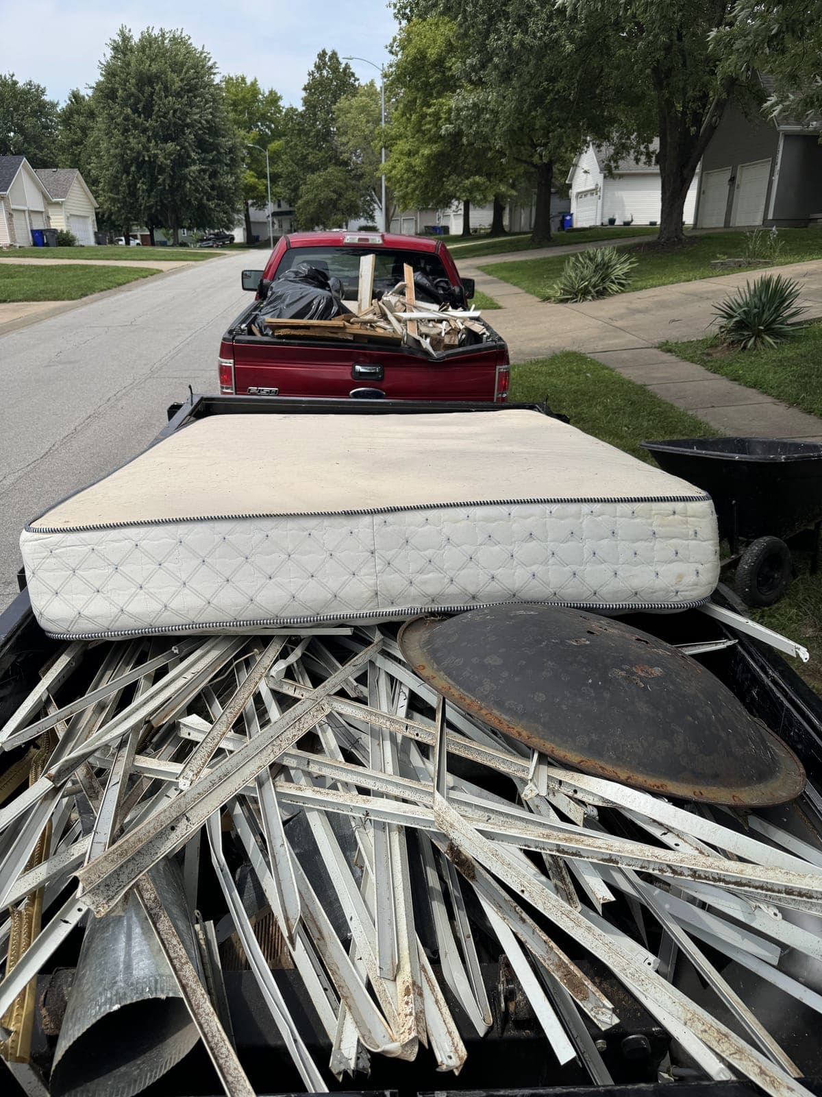 Dump trailer loaded curbside with a mattress on top of a full load of metal siding, framing pieces, and a round metal lid, with a loaded pickup truck ahead of it in the Kansas City metro