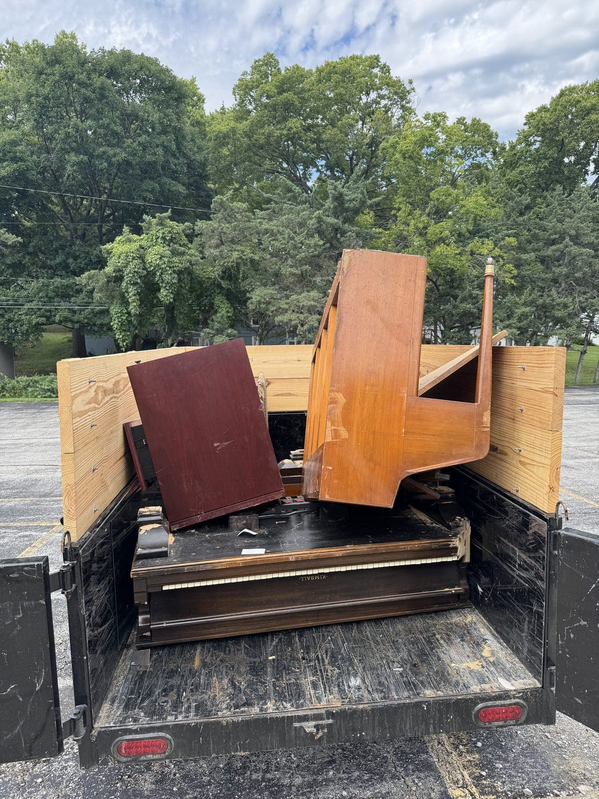 Dump trailer with raised wood side extensions loaded with an upright Kimball piano laid on its back with the keyboard exposed, alongside wood furniture panels and a bed rail, on a piano removal job in the Kansas City metro